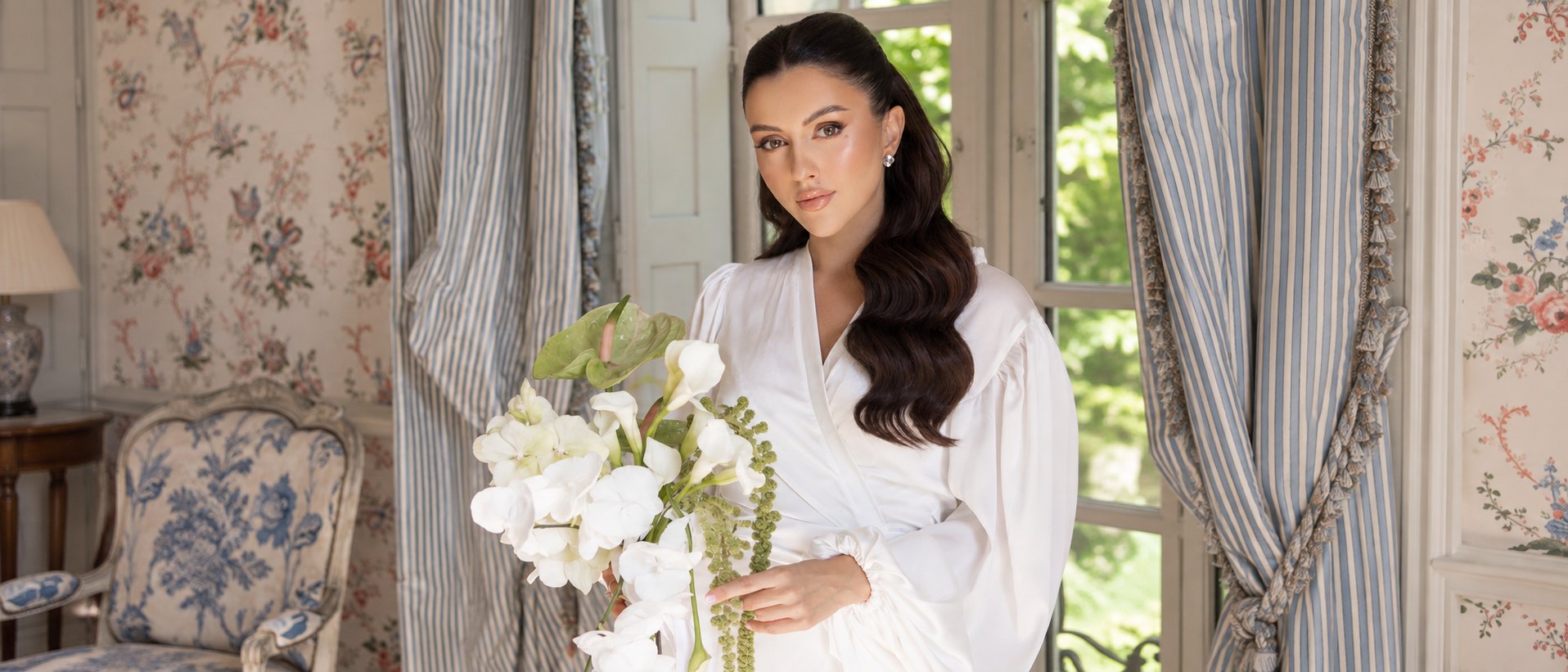 Mariée en peignoir de satin blanc, bouquet de fleurs blanches à la main, dans un salon aux rideaux rayés — maquillage signé Sanaa Make Up Académie